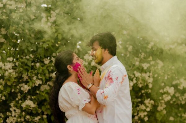 A couple enjoying a colorful pre-wedding shoot in Jaipur, playfully applying Holi colors to each other, surrounded by lush greenery and floral backdrops.