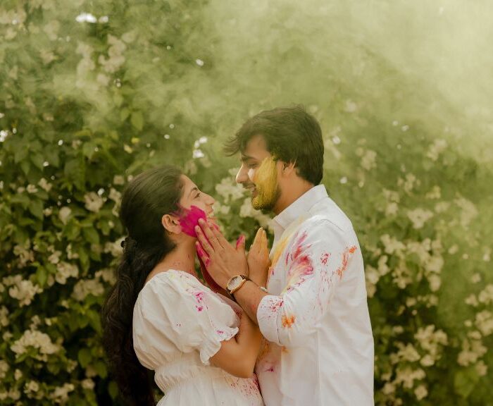 A couple enjoying a colorful pre-wedding shoot in Jaipur, playfully applying Holi colors to each other, surrounded by lush greenery and floral backdrops.