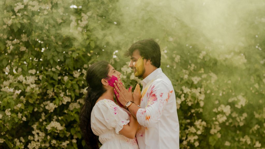 A couple enjoying a colorful pre-wedding shoot in Jaipur, playfully applying Holi colors to each other, surrounded by lush greenery and floral backdrops.