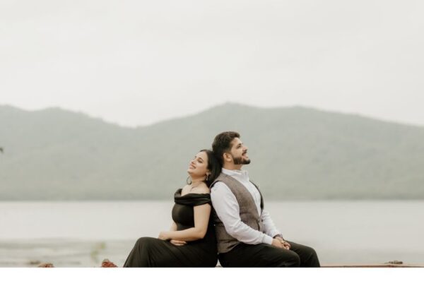 A couple sitting back-to-back, smiling peacefully during a pre-wedding shoot captured by a wedding photographer in Jaipur, with a stunning lake and mountains in the background.