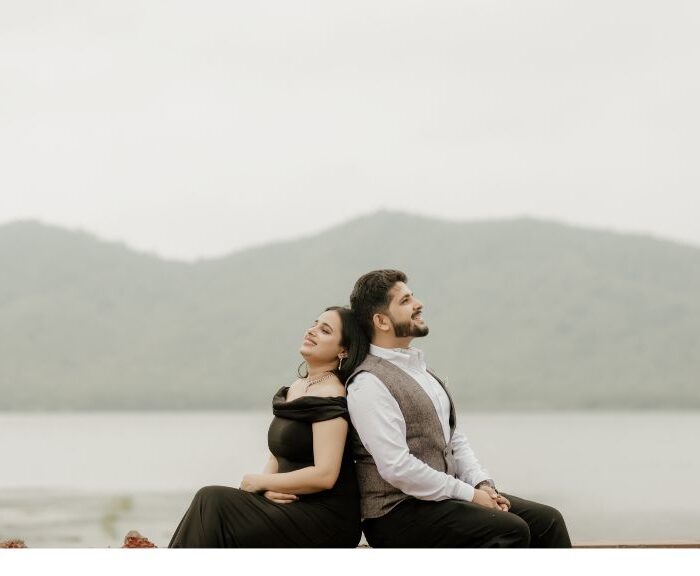 A couple sitting back-to-back, smiling peacefully during a pre-wedding shoot captured by a wedding photographer in Jaipur, with a stunning lake and mountains in the background.