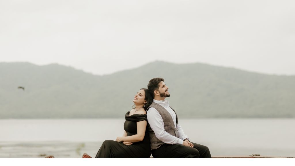 A couple sitting back-to-back, smiling peacefully during a pre-wedding shoot captured by a wedding photographer in Jaipur, with a stunning lake and mountains in the background.