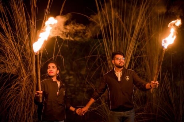 A couple holding torches during a pre-wedding shoot at night in a rustic setting with tall grass in the background.