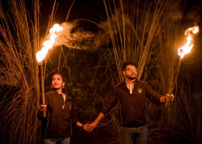 A couple holding torches during a pre-wedding shoot at night in a rustic setting with tall grass in the background.