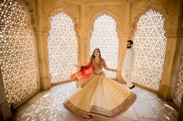 A couple dressed in traditional attire during a pre-wedding shoot in Jaipur, with the bride twirling in a lehenga and the groom walking behind her in a white suit.