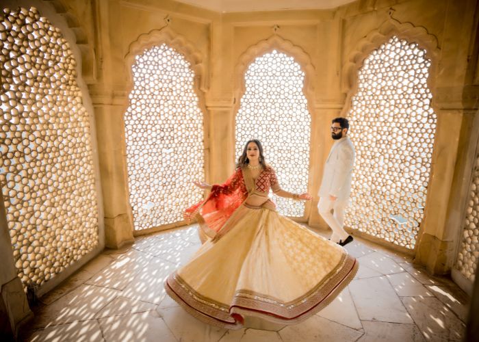 A couple dressed in traditional attire during a pre-wedding shoot in Jaipur, with the bride twirling in a lehenga and the groom walking behind her in a white suit.