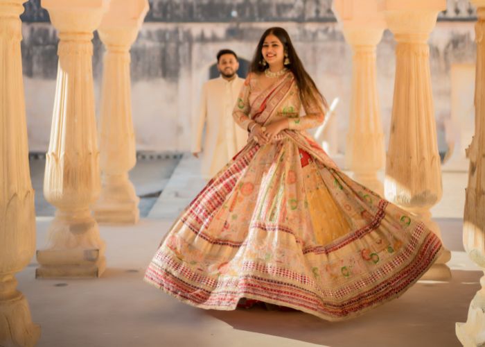 A couple during a pre-wedding shoot in Jaipur, with the bride twirling in a colorful lehenga, while the groom walks behind her in a cream-colored outfit.