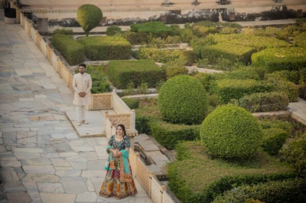 A pre-wedding couple photoshoot in a beautifully manicured garden, with the bride in a colorful lehenga and the groom in traditional attire standing in a serene and picturesque setting