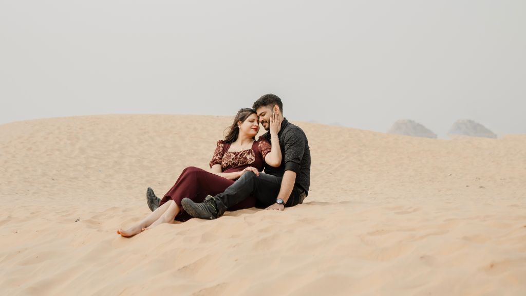 A couple enjoys a romantic moment in the desert during their pre-wedding shoot in Jaipur, sitting on the sand and laughing together.