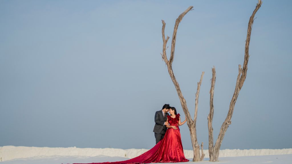 A romantic pre-wedding photoshoot featuring a couple in a stunning red gown and suit, standing together in a scenic location with a minimalist tree backdrop.