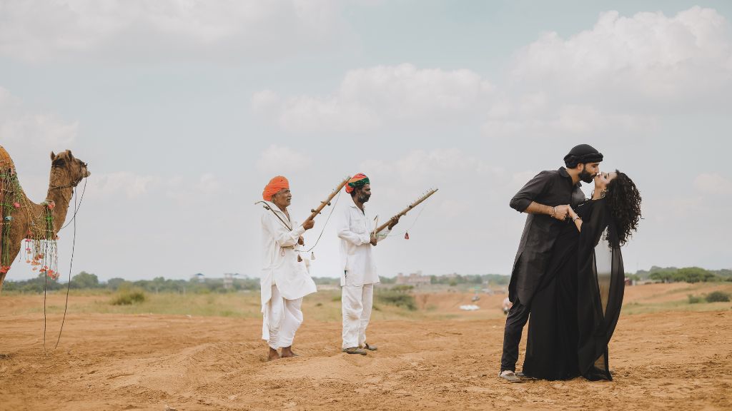 A beautiful pre-wedding shoot in Jaipur featuring a couple sharing a kiss while camel herders in traditional attire and a camel stand in the background.