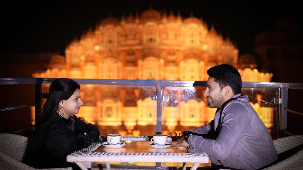 A couple enjoying a coffee together in a pre-wedding shoot in Jaipur, with the beautiful Hawa Mahal illuminated in the background.