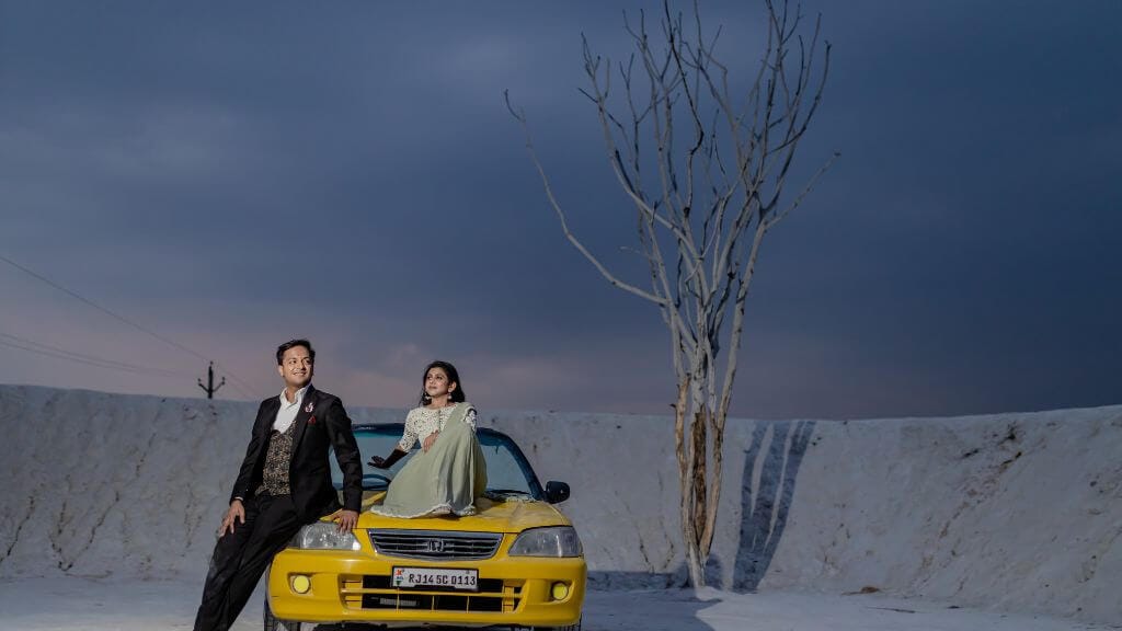 A couple posing on a yellow car in a pre-wedding shoot in Jaipur, with a minimalist tree and dramatic skies in the background.