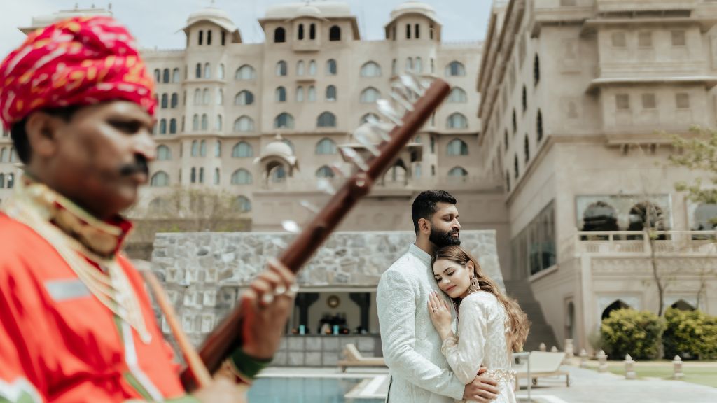 A couple embraces during a pre-wedding shoot in Jaipur, while a traditional musician plays the flute in the foreground.