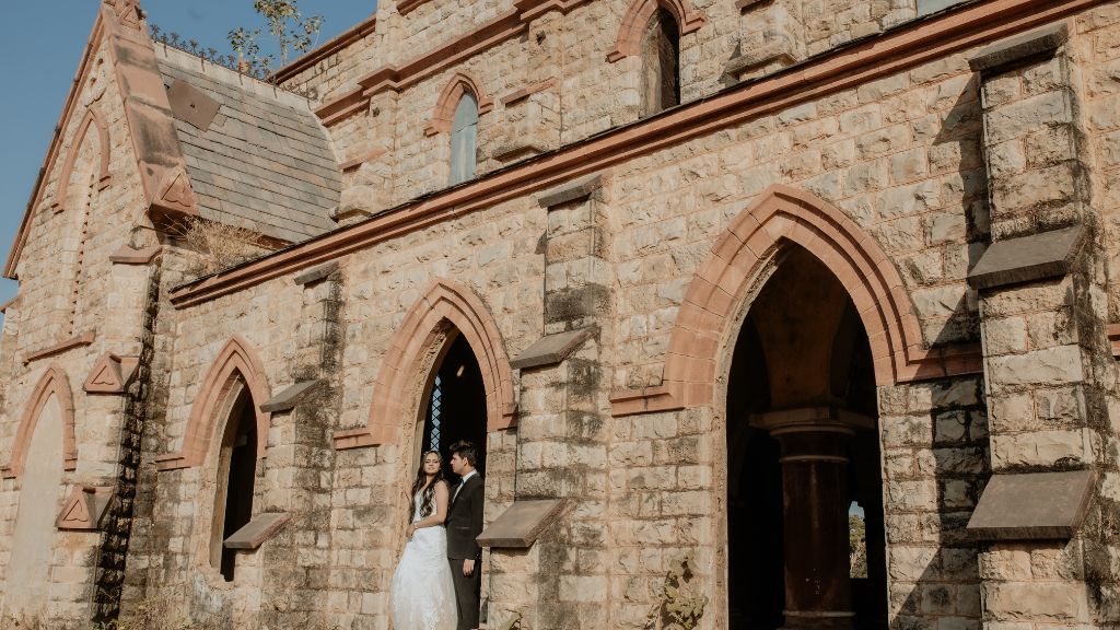 A couple poses for a pre-wedding photo in front of a historic stone building in Jaipur, with the bride in a white gown and the groom in a black suit.