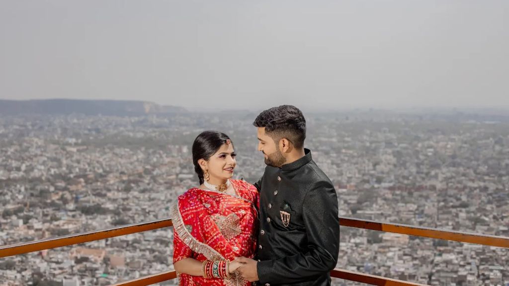 A couple stands together in a pre-wedding shoot, overlooking the city of Jaipur with the bride in a red traditional outfit and the groom in a black sherwani.