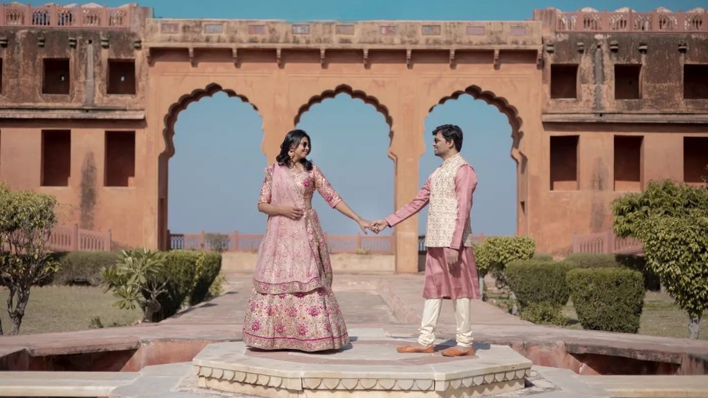 A couple holds hands in a pre-wedding shoot, with the bride in a pink traditional outfit and the groom in a matching attire, standing in front of a beautiful historical archway in Jaipur.