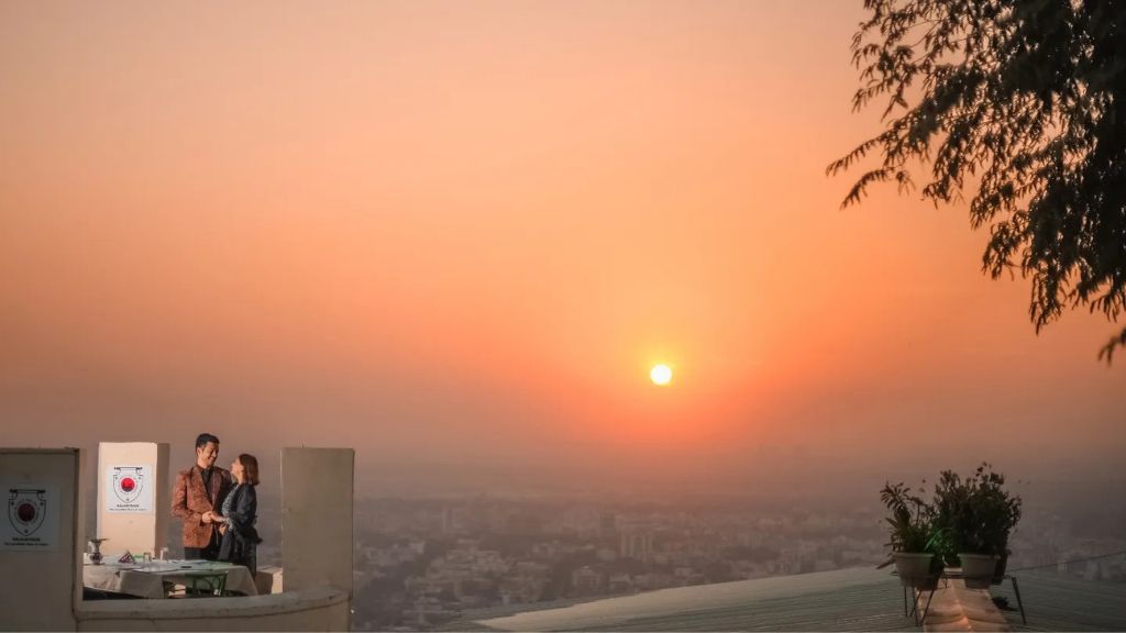 A couple stands on a rooftop during a pre-wedding shoot in Jaipur, with the sunset casting a warm glow over the city in the background.