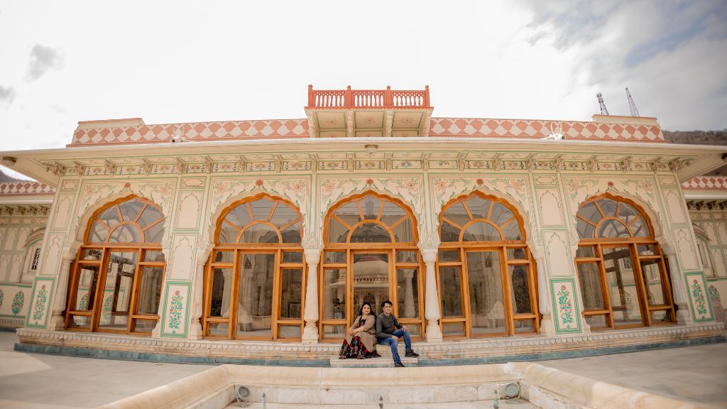 A couple sitting on the ground in front of a beautifully designed palace with intricate arches and windows during a pre-wedding photoshoot in Jaipur.