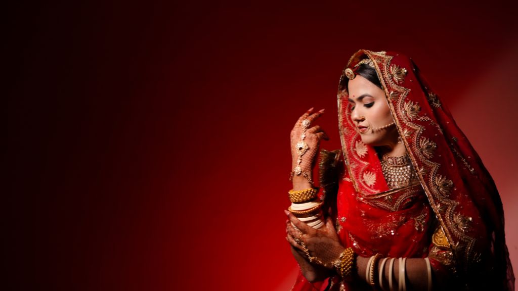 A bride in a red and gold embroidered lehenga poses gracefully against a deep red background, showcasing traditional Indian bridal beautyBest Wedding Photographers in Jaipur.