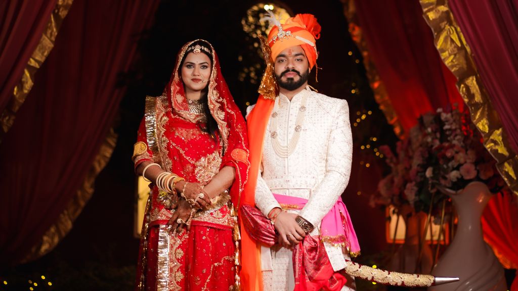  An Indian bride in a red and gold lehenga and a groom in an ivory sherwani with a turban pose elegantly in a royal wedding setting.