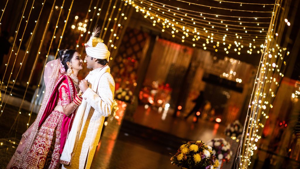 An Indian bride in a red and gold lehenga and a groom in an ivory sherwani share a romantic moment under fairy lights at their wedding venue.