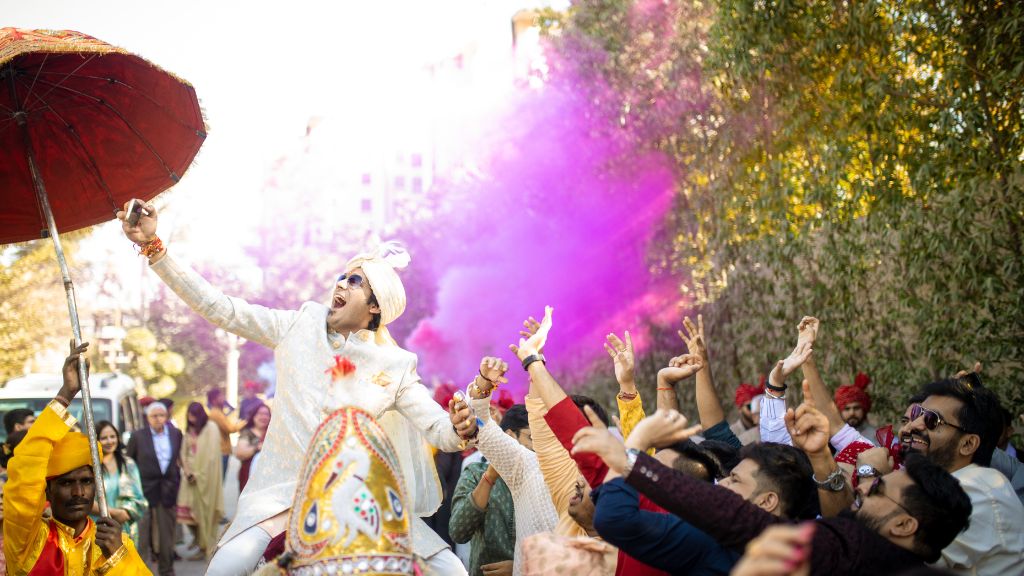 An Indian groom in a white sherwani and turban dances joyfully on a decorated horse, surrounded by friends and family with vibrant purple smoke in the background.