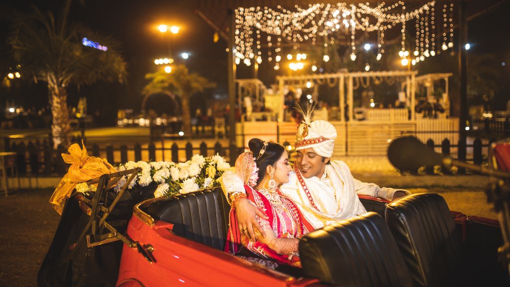 An Indian bride and groom sit closely in a vintage red car, sharing a romantic moment under twinkling lights at their wedding venue.