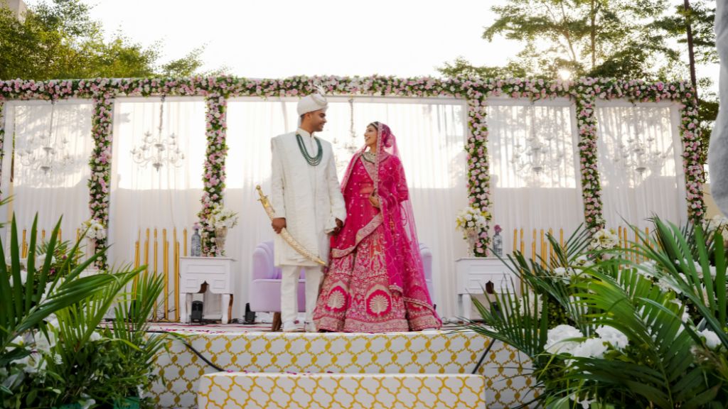 An Indian bride in a pink lehenga and groom in an ivory sherwani stand on a floral-decorated wedding stage, looking lovingly at each other.