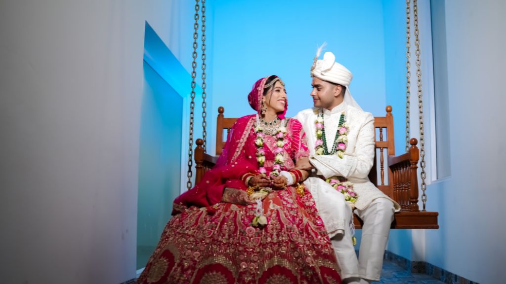 A newlywed Indian couple sits on a wooden swing, dressed in traditional wedding attire, smiling lovingly at each other.