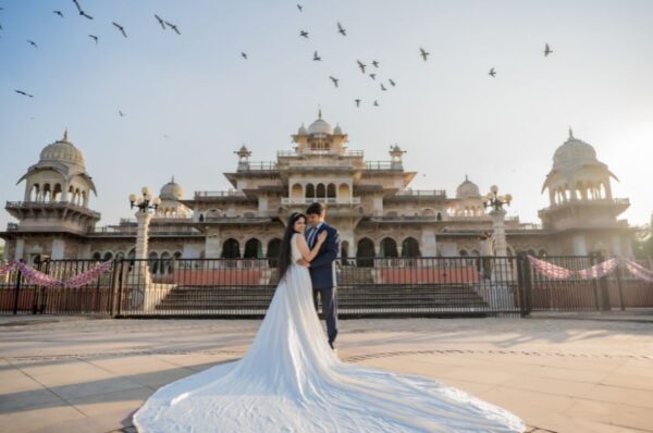 A couple in wedding attire poses for a pre-wedding photoshoot in Jaipur, standing in front of a majestic palace with a flowing bridal gown, golden sunset, and birds flying in the sky.