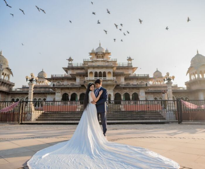 A couple in wedding attire poses for a pre-wedding photoshoot in Jaipur, standing in front of a majestic palace with a flowing bridal gown, golden sunset, and birds flying in the sky.