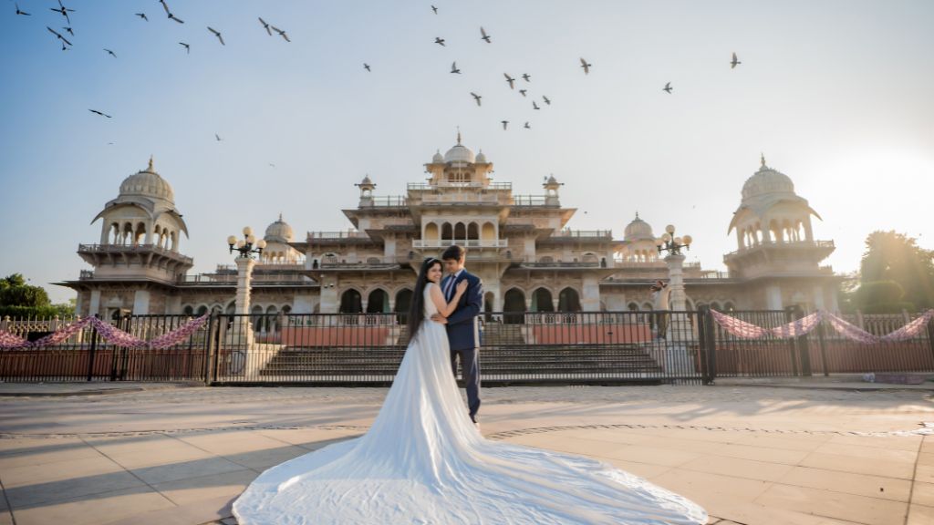 A couple in wedding attire poses for a pre-wedding photoshoot in Jaipur, standing in front of a majestic palace with a flowing bridal gown, golden sunset, and birds flying in the sky.