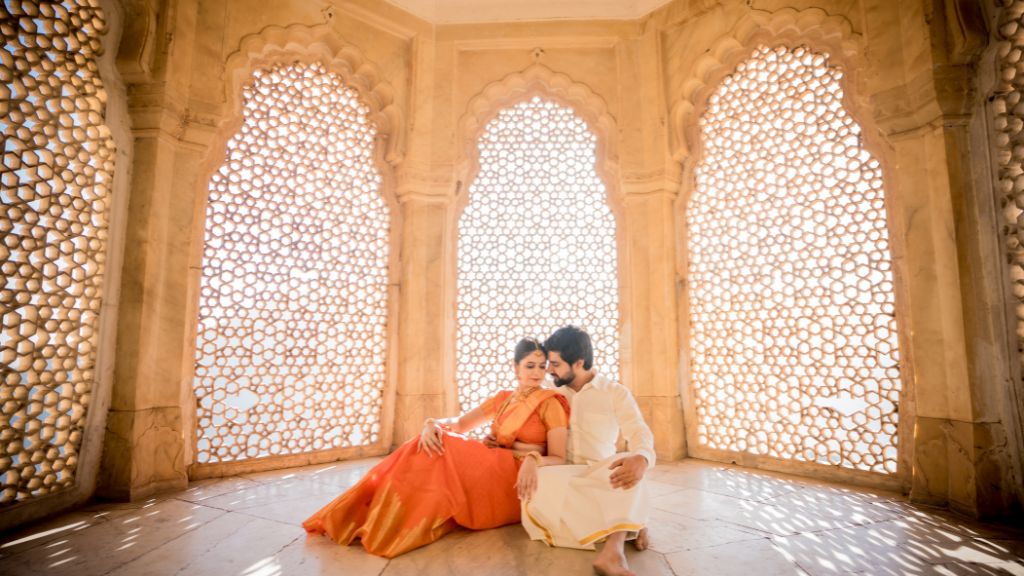 A couple dressed in traditional Indian attire sits in an elegant palace with intricate lattice windows, bathed in warm golden sunlight during a pre-wedding photoshoot in Jaipur.