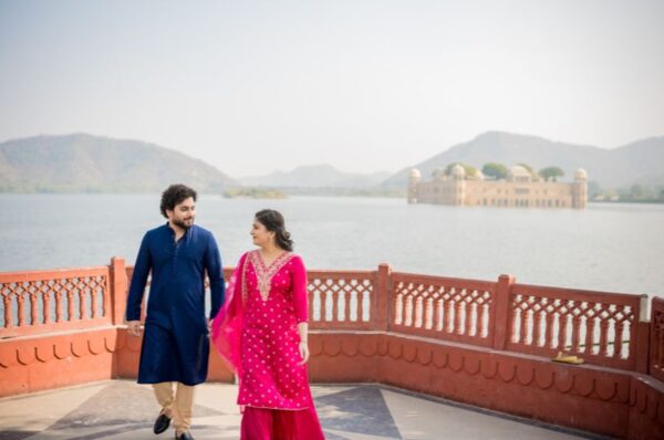 A couple in traditional attire walking together during a pre-wedding photoshoot with a scenic lake and mountains in the background.