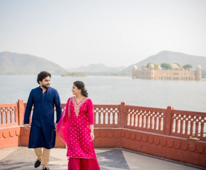 A couple in traditional attire walking together during a pre-wedding photoshoot with a scenic lake and mountains in the background.