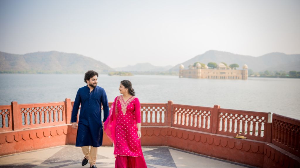 A couple in traditional attire walking together during a pre-wedding photoshoot with a scenic lake and mountains in the background.