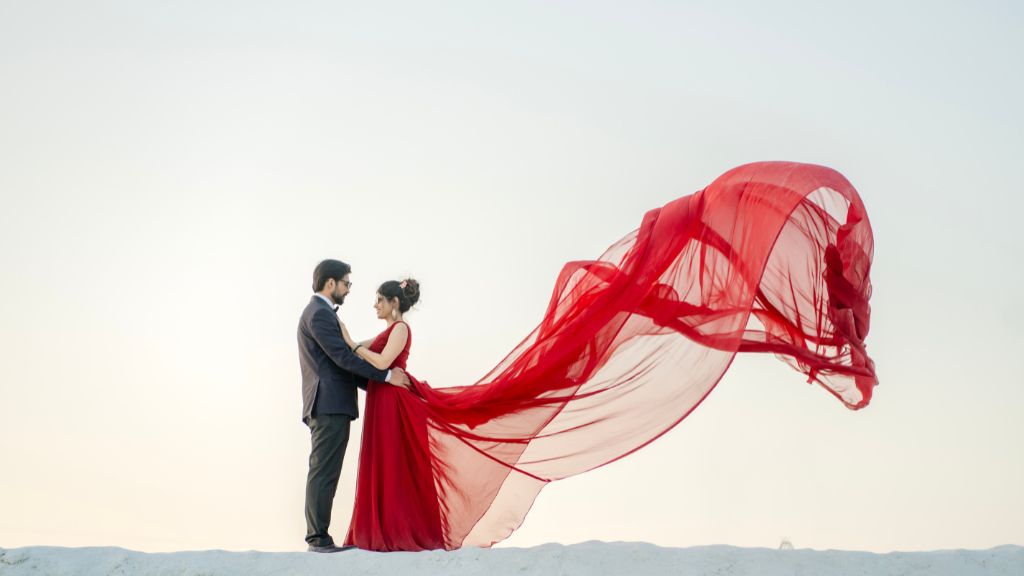 A couple in elegant attire poses for a pre-wedding photoshoot, with the bride’s flowing red fabric captured in the wind