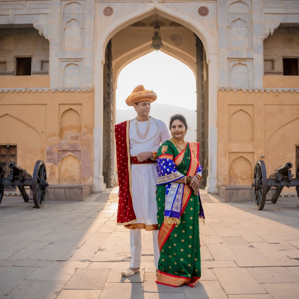 Couple in traditional Indian attire, man in a royal white outfit with a turban and woman in a green and red saree, standing in front of an ancient fort gate in Jaipur during a post-wedding photoshoot.