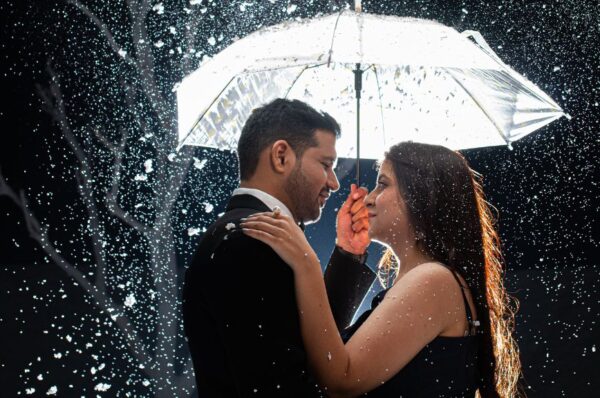 A couple embraces under an umbrella during a pre-wedding shoot in Jaipur, with snow-like sparkles falling around them, creating a romantic atmosphere.