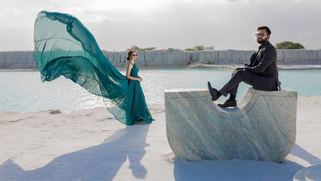 Couple posing by a serene turquoise lake, woman in a flowing gown and man in a formal suit during a pre-wedding shoot near Jaipur