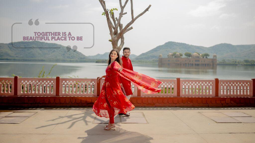 A joyful couple in matching red outfits posing near Jal Mahal lake in Jaipur, with the quote “Together is a beautiful place to be” displayed above them in a romantic wedding-themed image.