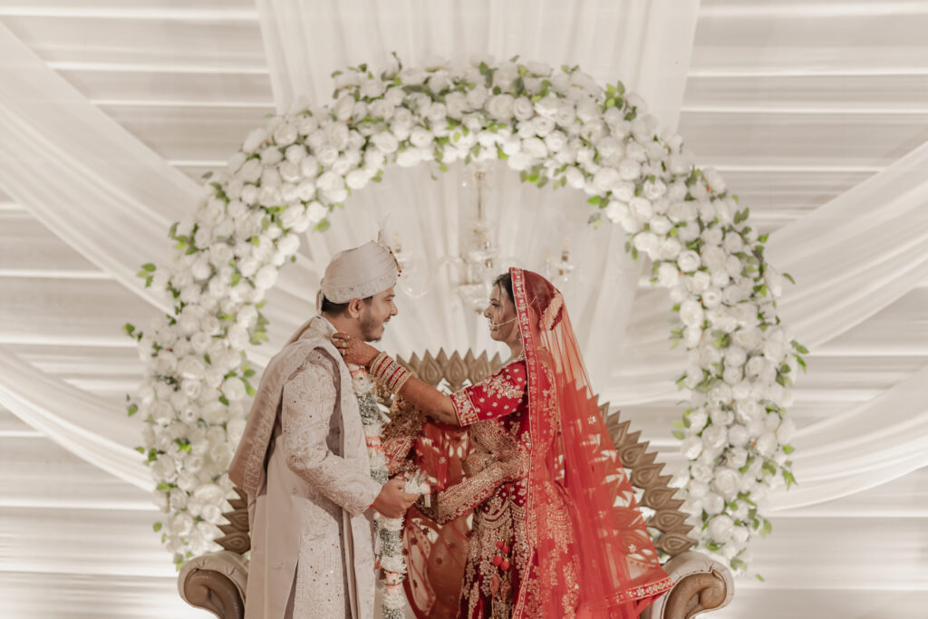 An Indian bride in a red lehenga and groom in a cream sherwani sharing a sweet, romantic moment on a stage under a white floral arch, captured by Wedding Photographers in Jaipur.