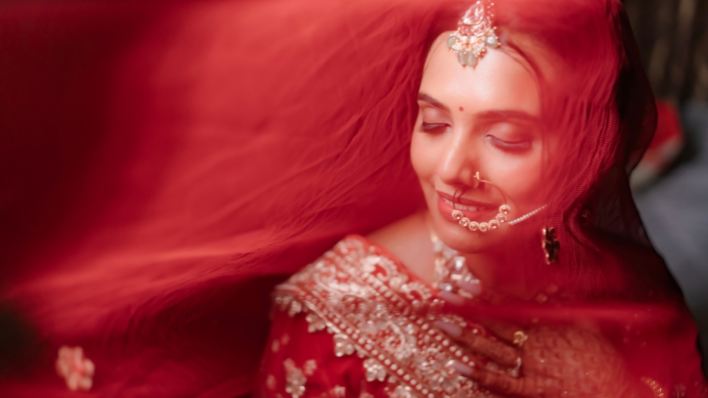 A close-up of a smiling Indian bride draped under a sheer red veil, wearing traditional pearl and gold jewelry, captured by Wedding Photographers in Jaipur.