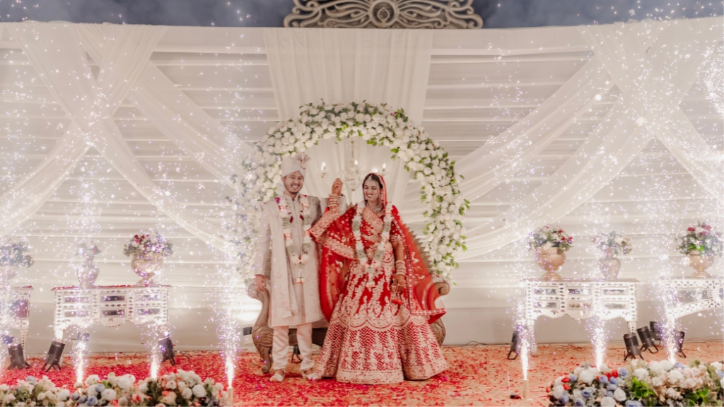 A joyful Indian bride and groom holding hands on a stage decorated with white drapery, a floral arch, and bright sparkler fireworks, photographed by Wedding Photographers in Jaipur.