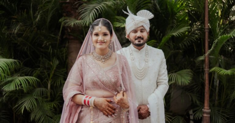 An elegant portrait of an Indian bride in a soft pink lehenga and groom in an ivory sherwani standing against lush green foliage, captured by Wedding Photographers in Jaipur.