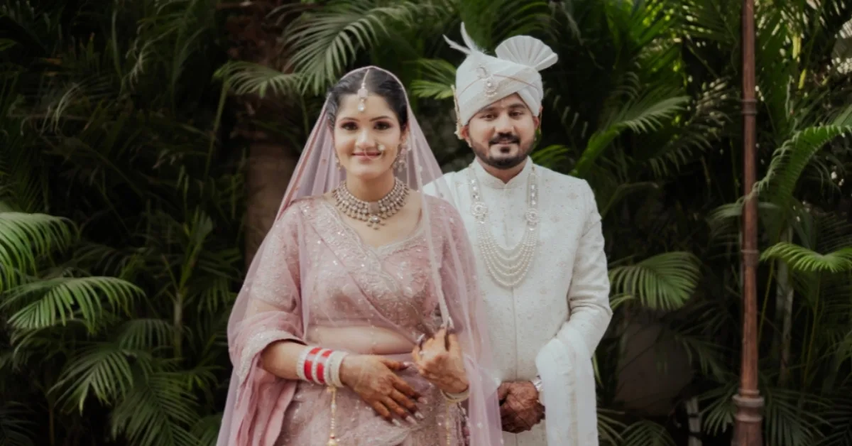 An elegant portrait of an Indian bride in a soft pink lehenga and groom in an ivory sherwani standing against lush green foliage, captured by Wedding Photographers in Jaipur.