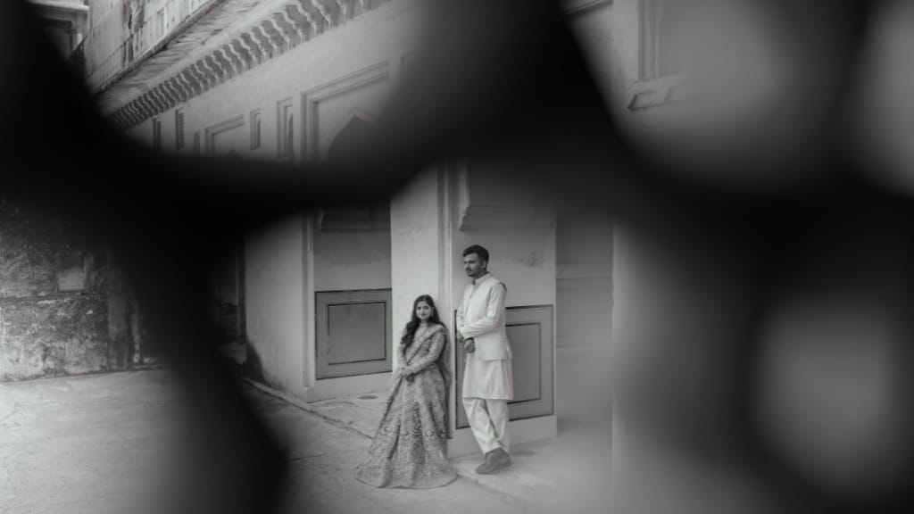 Couple posing in traditional attire for a pre wedding photoshoot at a heritage palace, framed through a stone lattice in black and white.