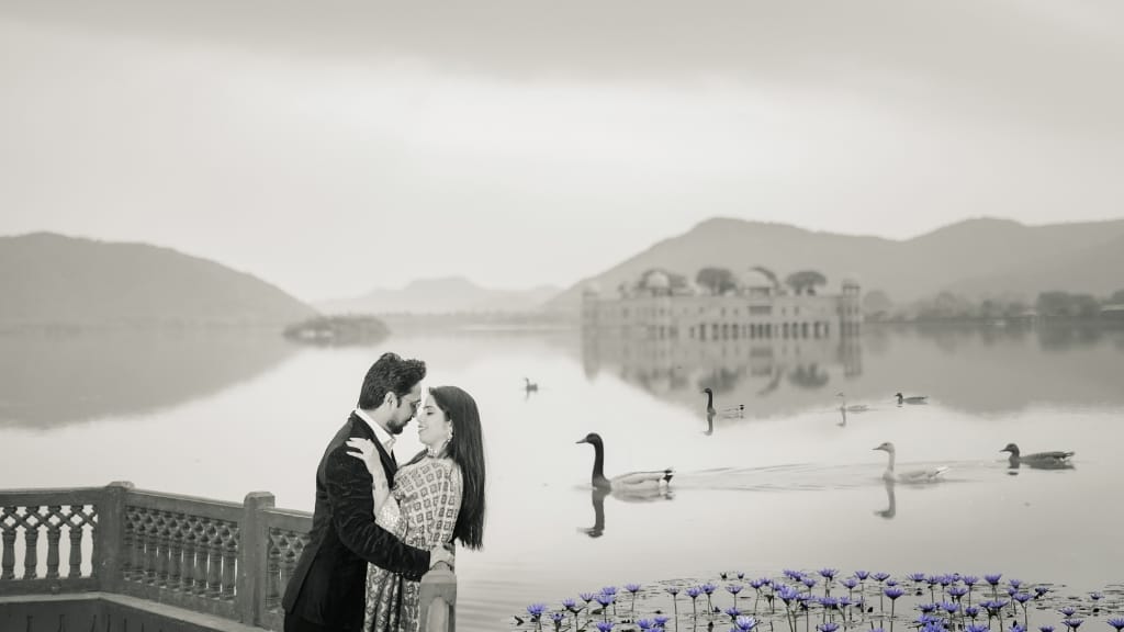 Couple embracing during a pre wedding photoshoot at Jal Mahal, Jaipur, featuring a lake with ducks and purple lotus flowers in the foreground.