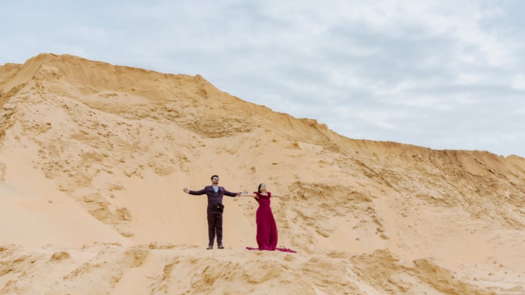 A couple posing with open arms in a desert landscape for their pre wedding photoshoot, wearing a maroon gown and a formal suit against golden sand dunes.